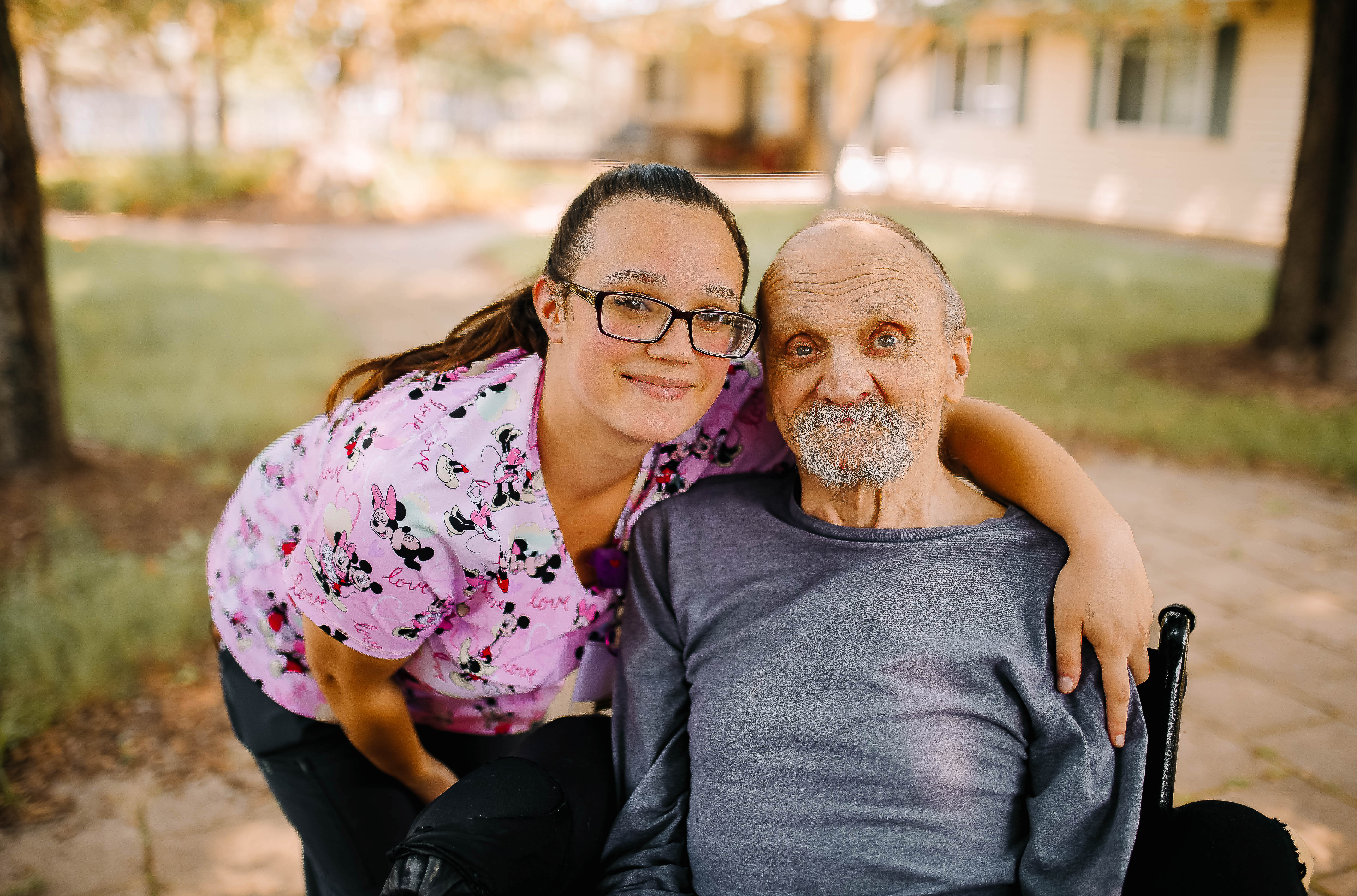 A woman in glasses embraces an elderly man while seated in her wheelchair, showcasing a moment of warmth and connection