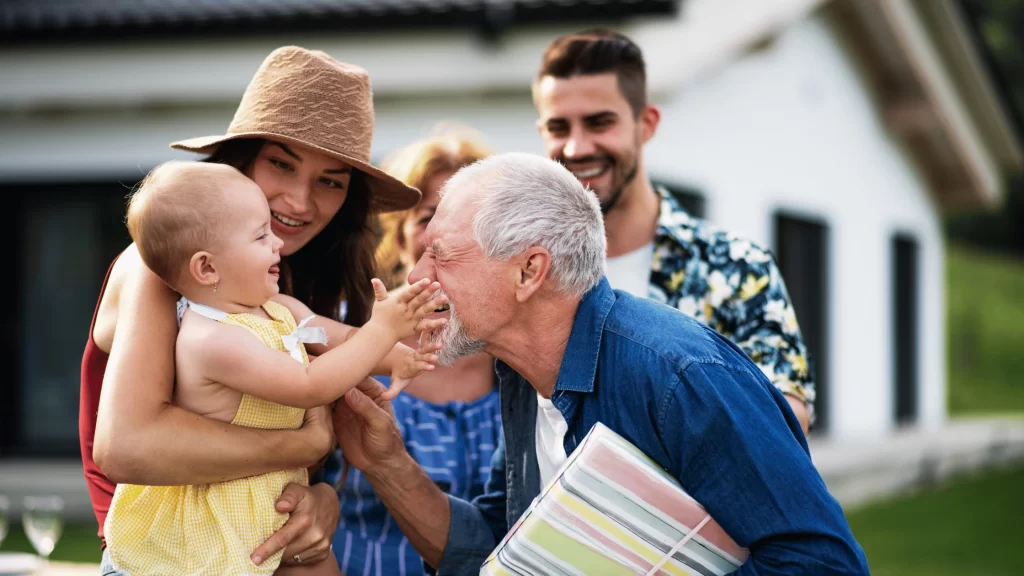 A family visits their senior family member outside an assisted living home, holding a baby and sharing a moment together