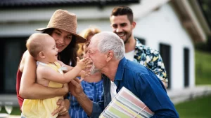 A family visits their senior family member outside an assisted living home, holding a baby and sharing a moment together