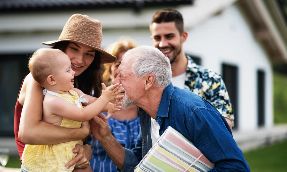 A family visits their senior family member outside an assisted living home, holding a baby and sharing a moment together